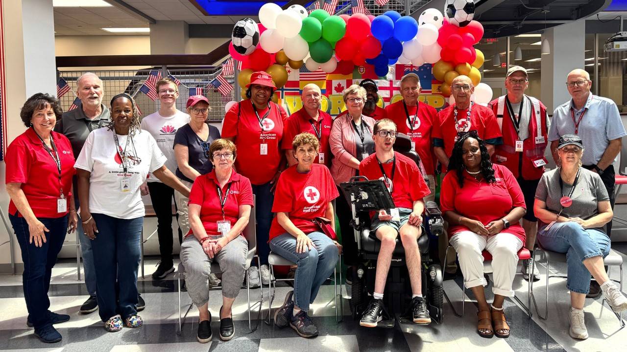 A large group of about fifteen smiling American Red Cross volunteers and staff pose together indoors for a group photo. Most are wearing bright red Red Cross T-shirts, while a few wear polo shirts or casual attire. They are gathered in a brightly lit hallway or atrium decorated with festive red, white, and blue balloon arches and hanging international flags in the background.