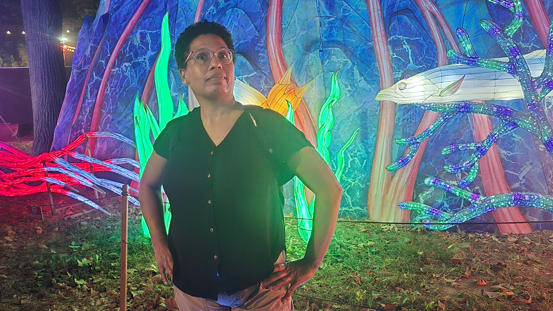 African American woman wearing a black shirt and tan pants stands with her hands on her hips in front of a lit backdrop that looks like an aquarium. 