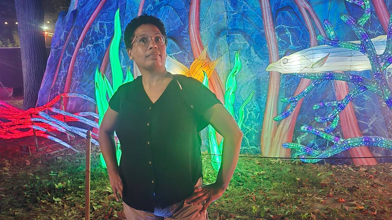 African American woman wearing a black shirt and tan pants stands with her hands on her hips in front of a lit backdrop that looks like an aquarium. 