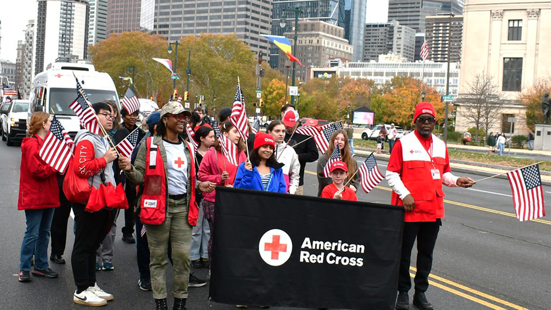 30 Red Cross volunteers wearing red and holding a banner walk in a parade.