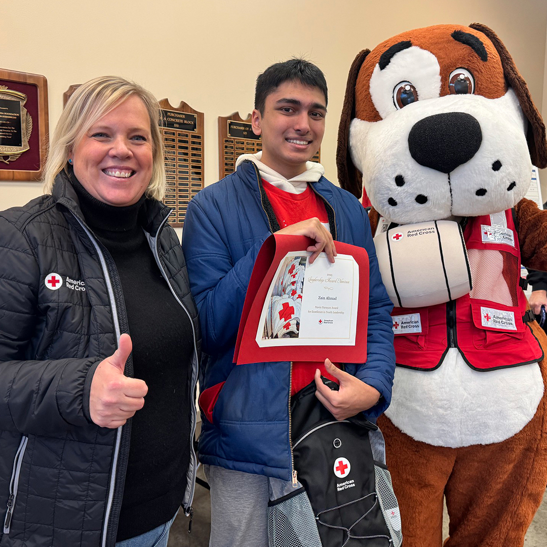 Zain Ahmad holds a certificate and Red Cross backpack. Jennifer Graham is pictured giving a thumbs up on his left and Red Cross mascot (person inside large dog costume wearing a Red Cross vest) is on his left. 