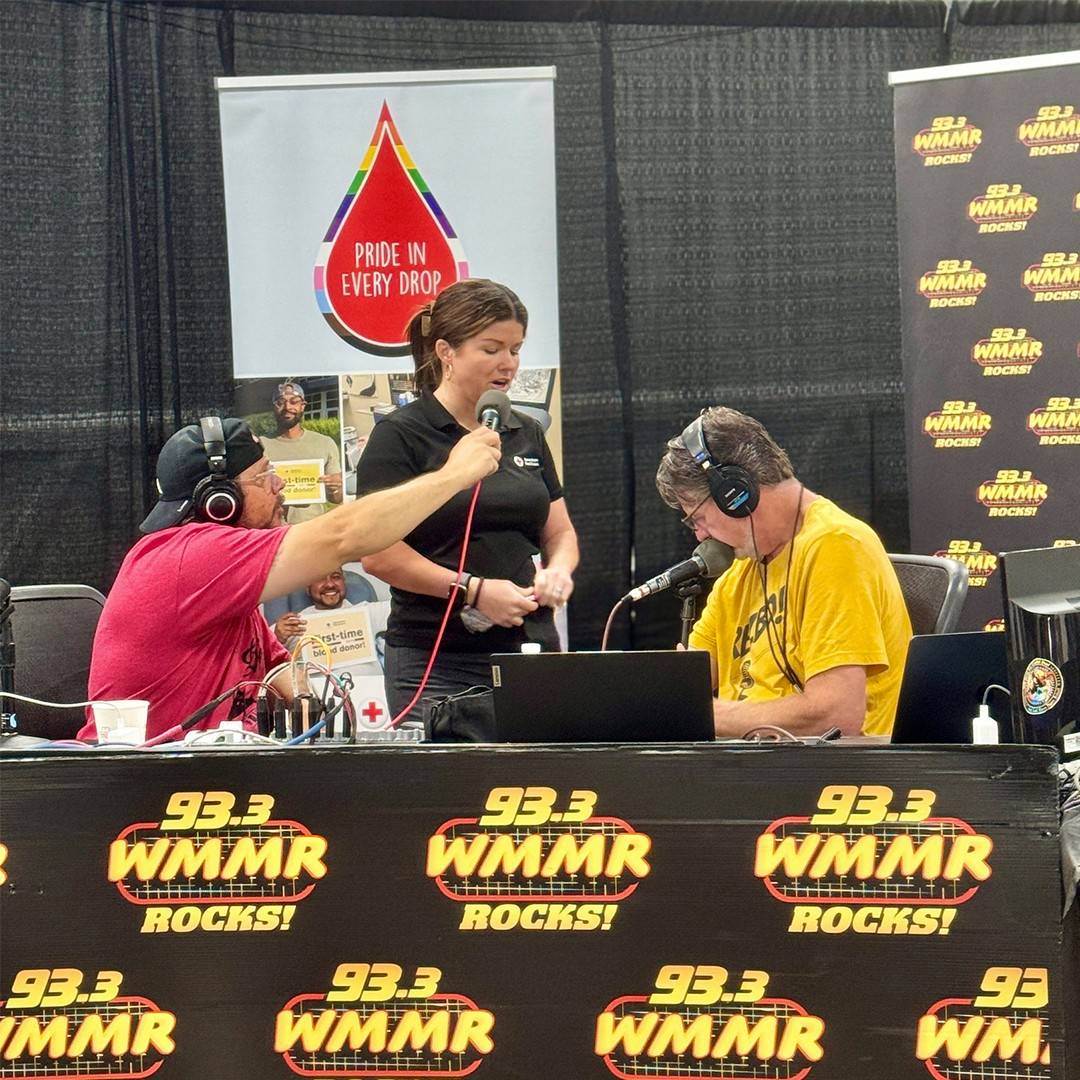 Blood Collections Supervisor Ashley Barrett, wearing a dark Red Cross uniform shirt and a headset mic, checks host Preston Elliot's arm to locate his veins. Preston, wearing a red t-shirt, baseball cap, and headphones, looks on while morning show producer Casey Boy stands nearby holding a handheld microphone during the live broadcast. Multiple signs featuring the "93.3 WMMR ROCKS!" logo are visible at the front of the broadcast table.