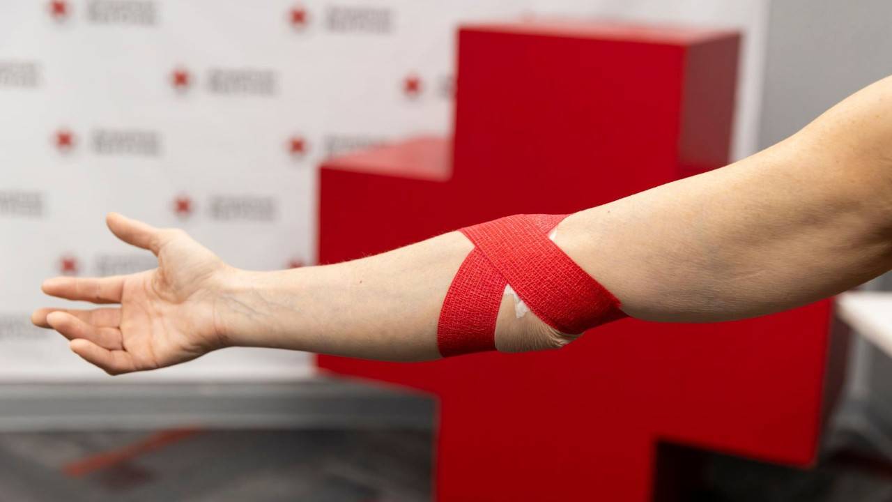 A close-up view of a person's bare arm resting on a table, with a red elastic band tied around the upper arm in preparation for a blood donation.