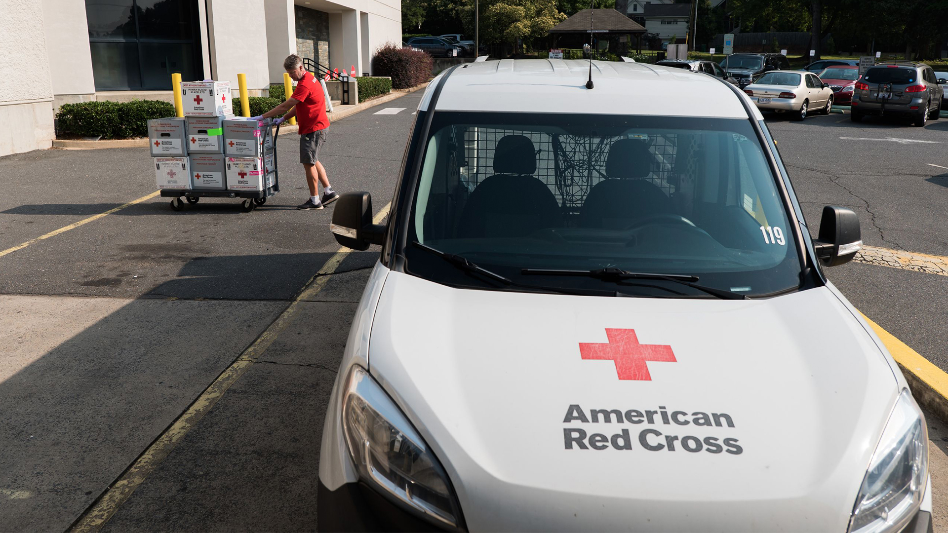 A Red Cross volunteer in a red shirt pushes a metal cart stacked  with boxes in a parking lot. In the foreground, the front hood of a white American Red Cross delivery vehicle is prominently visible.
