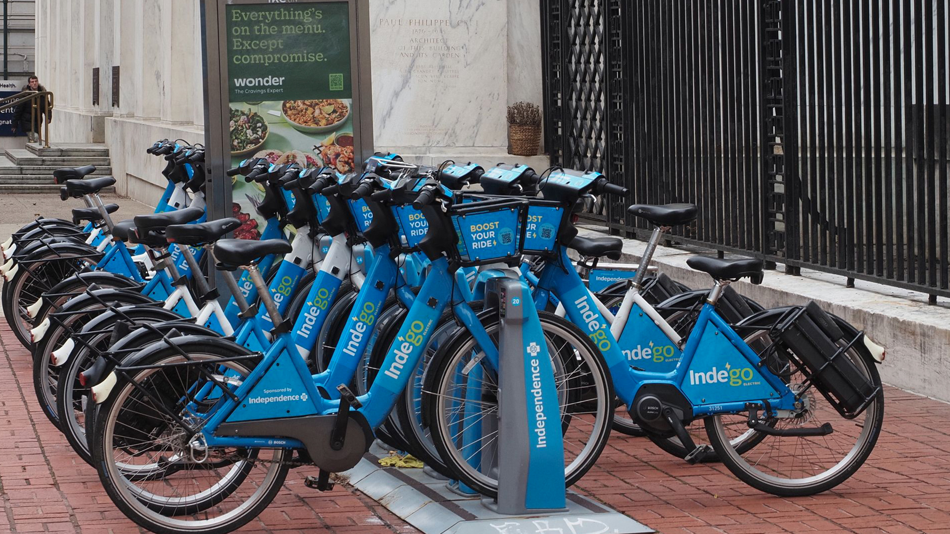 A row of blue Indego electric bicycles parked in a charging dock along a sidewalk at 10th and Chestnut streets in Philadelphia, with an automated charging station kiosk visible in the background.