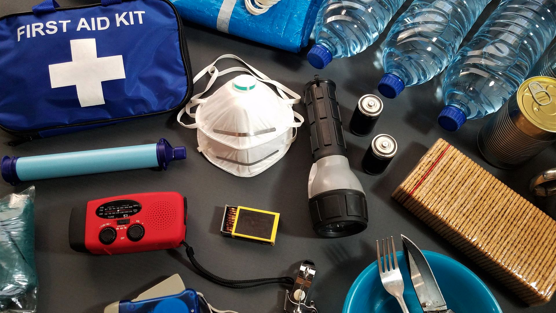 An overhead view of various emergency kit supplies laid out on a dark surface, including a blue first aid bag with a white cross, water bottles, a flashlight, extra batteries, a red emergency radio, canned food, eating utensils, a white face mask, a blue emergency blanket, and matches.