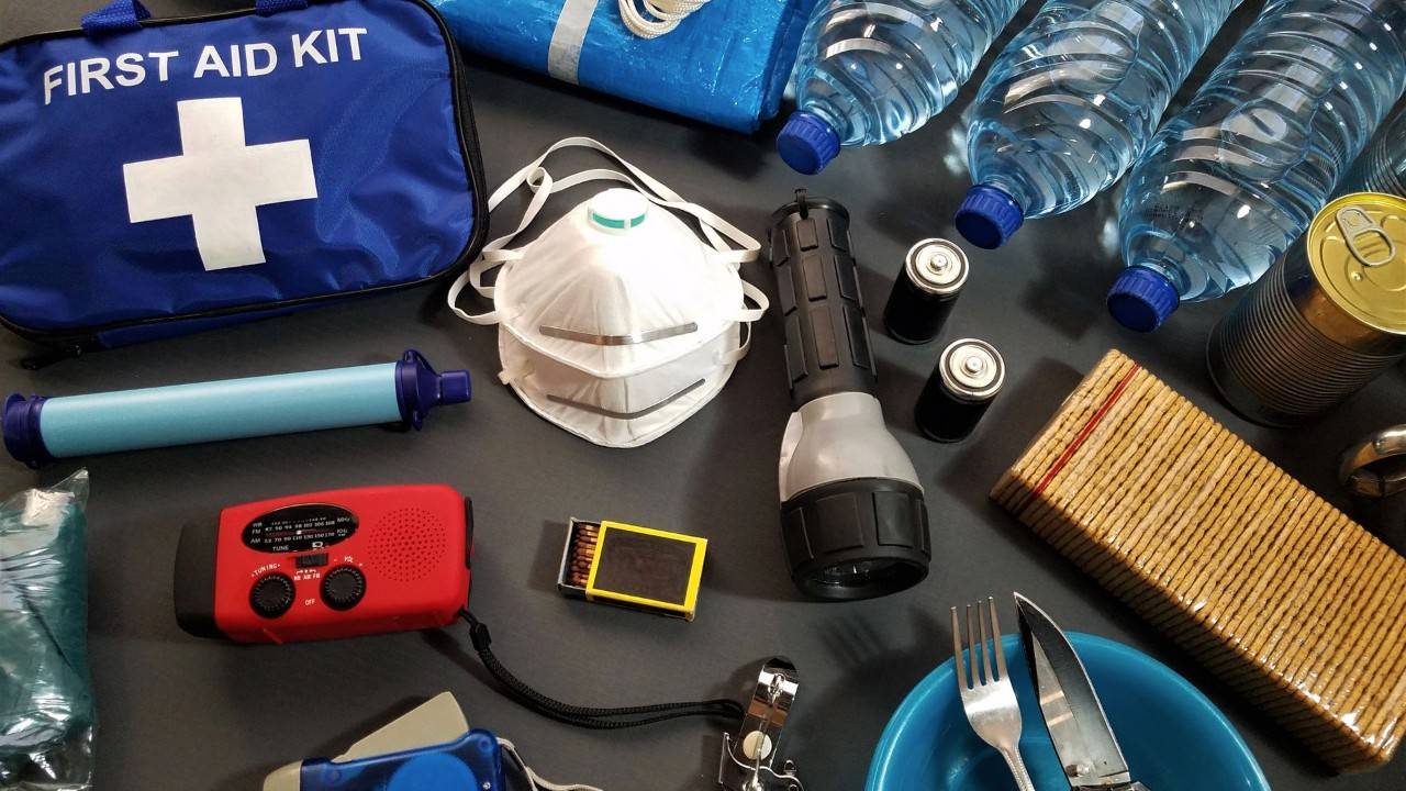 An overhead view of various emergency kit supplies laid out on a dark surface, including a blue first aid bag with a white cross, water bottles, a flashlight, extra batteries, a red emergency radio, canned food, eating utensils, a white face mask, a blue emergency blanket, and matches.