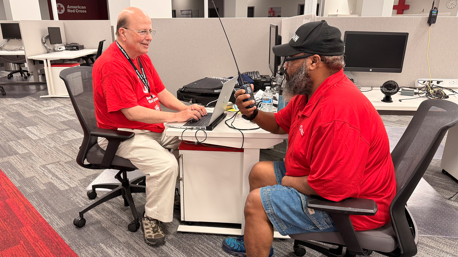 Two male Red Cross volunteers in red t-shirts sit at a workspace desk; one types on a laptop while the other operates a handheld amateur radio.