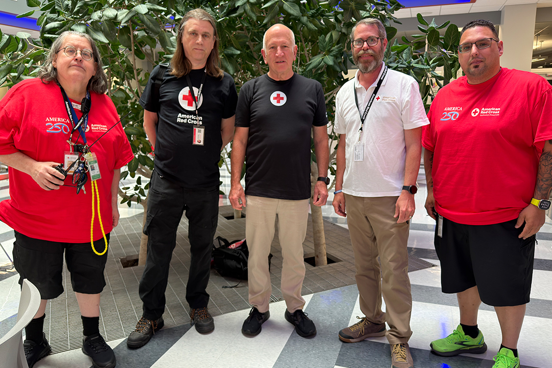 Five smiling American Red Cross volunteers stand side-by-side in a brightly lit building atrium with large green plants in the background. They are wearing Red Cross t-shirts, credentials around their necks, and portable amateur radios clipped to their belts or held in hand.