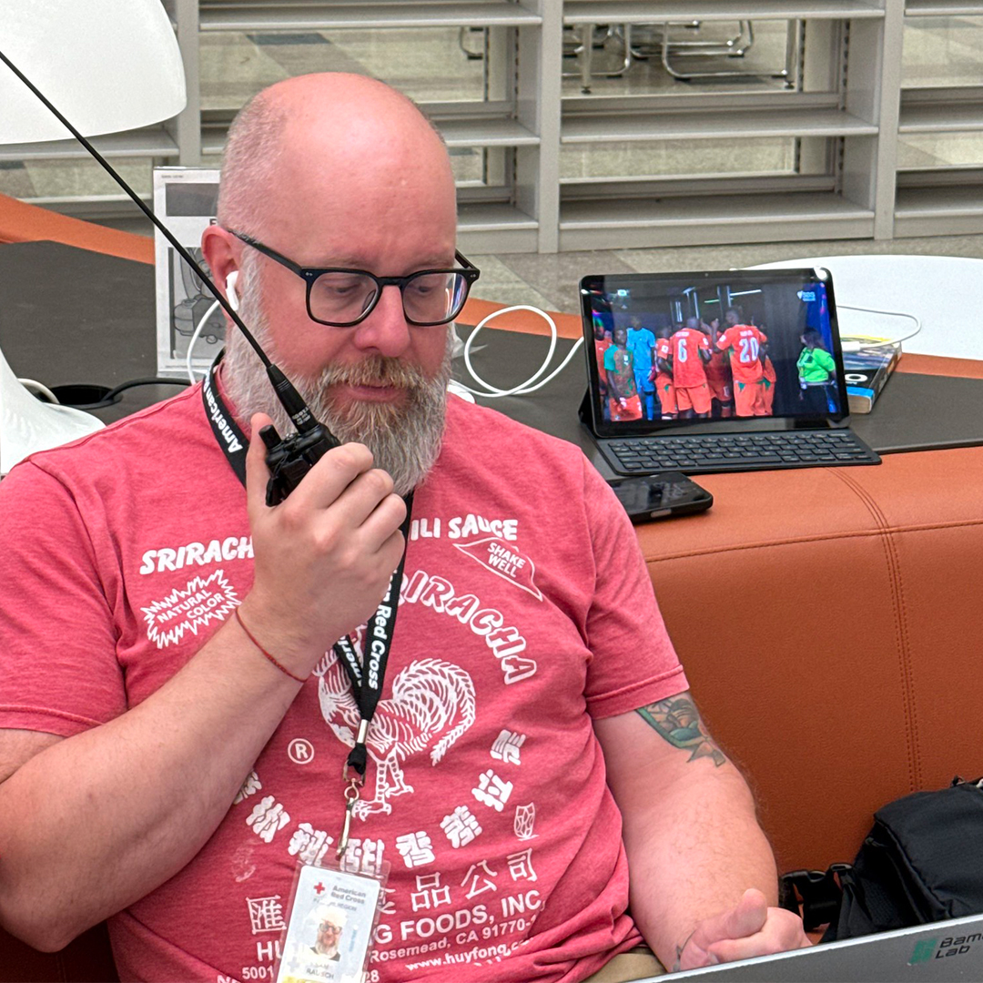 A close-up photograph of a bald man with a beard and glasses, wearing a pink Sriracha t-shirt and an ID badge, speaking into a handheld amateur radio.