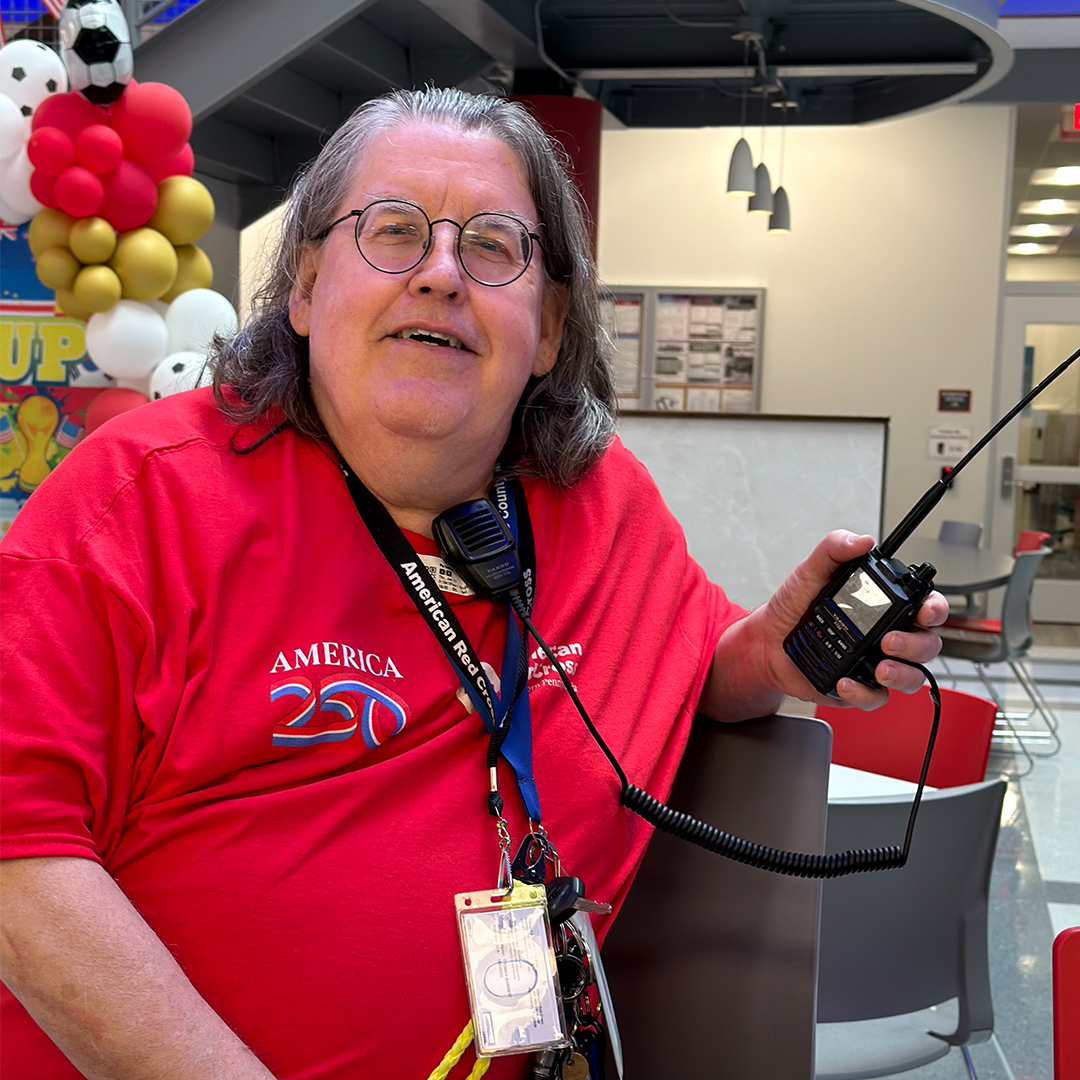 A close-up photograph of a smiling, middle-aged man with glasses and long grey hair, wearing a red American Red Cross "America 250" t-shirt. He has an official volunteer credential lanyard around his neck, a speaker microphone clipped near his collar, and is holding a handheld amateur (ham) radio with a long antenna in his left hand while seated indoors.