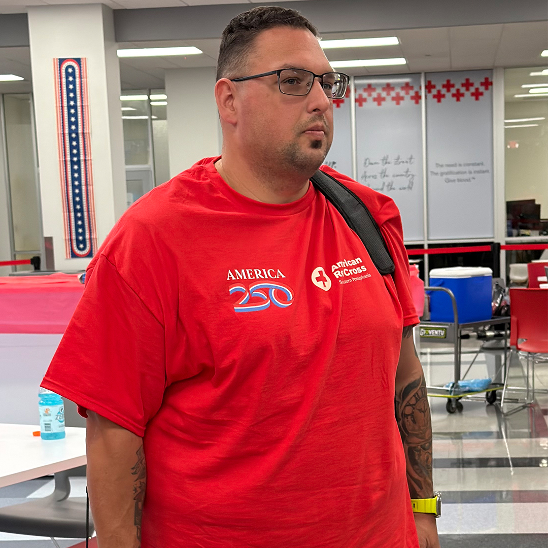 A close-up photograph of a man with short dark hair, glasses, and arm tattoos, wearing a red American Red Cross "America 250" t-shirt and a black backpack strap over his shoulder.