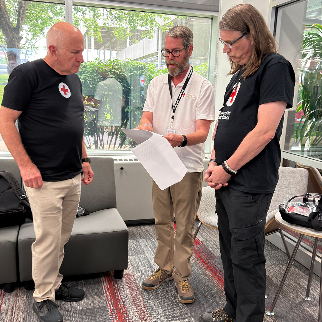 Three men stand indoors in a workspace reviewing a document together. Two of them wear black t-shirts with a red cross logo, and the middle man wears a white polo shirt with a lanyard.