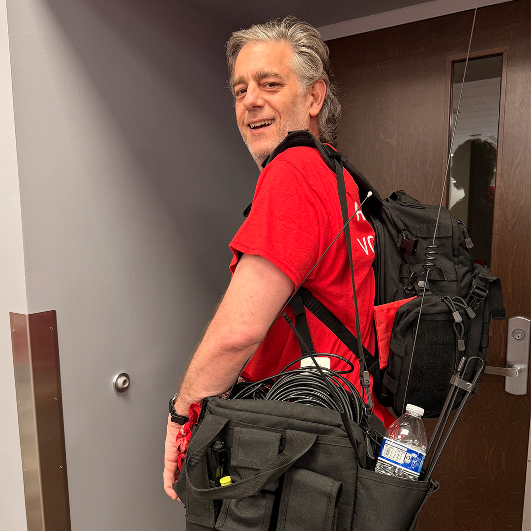 A close-up photograph of a smiling, middle-aged man with grey hair and a red American Red Cross t-shirt, carrying a black backpack and a large black gear bag filled with cables and equipment.