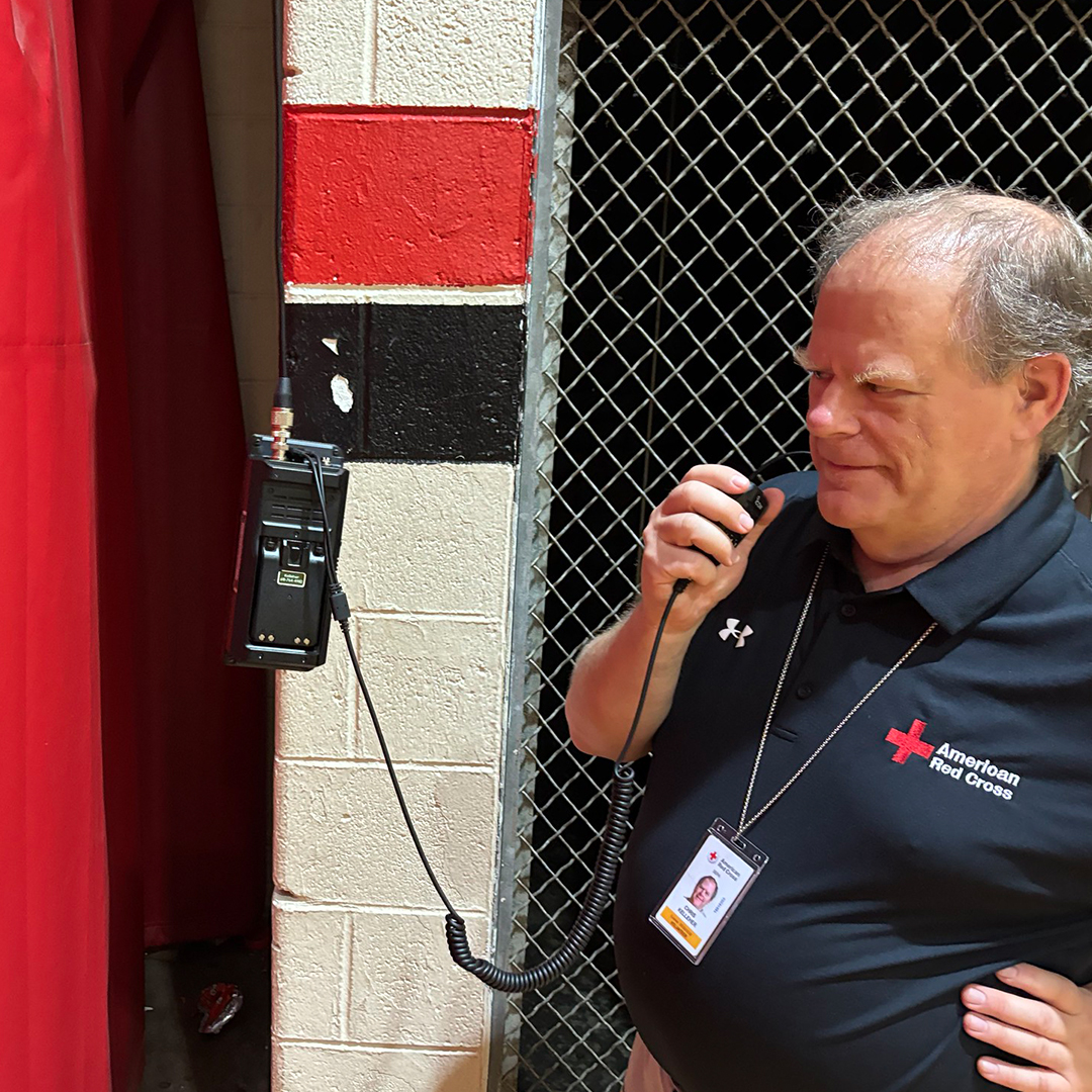 A close-up photograph of a smiling man with a black American Red Cross polo shirt and an ID badge, speaking into a handheld amateur radio with a red wall in the background.