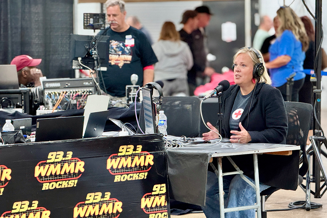 Jennifer Graham, CEO of the American Red Cross Southeastern Pennsylvania Region, wears a black blazer with a Red Cross logo over a white shirt and headphones. She sits at a broadcast table with microphones, gesturing with her hands while speaking on-air. In front of her are several signs featuring the "93.3 WMMR ROCKS!" logo.