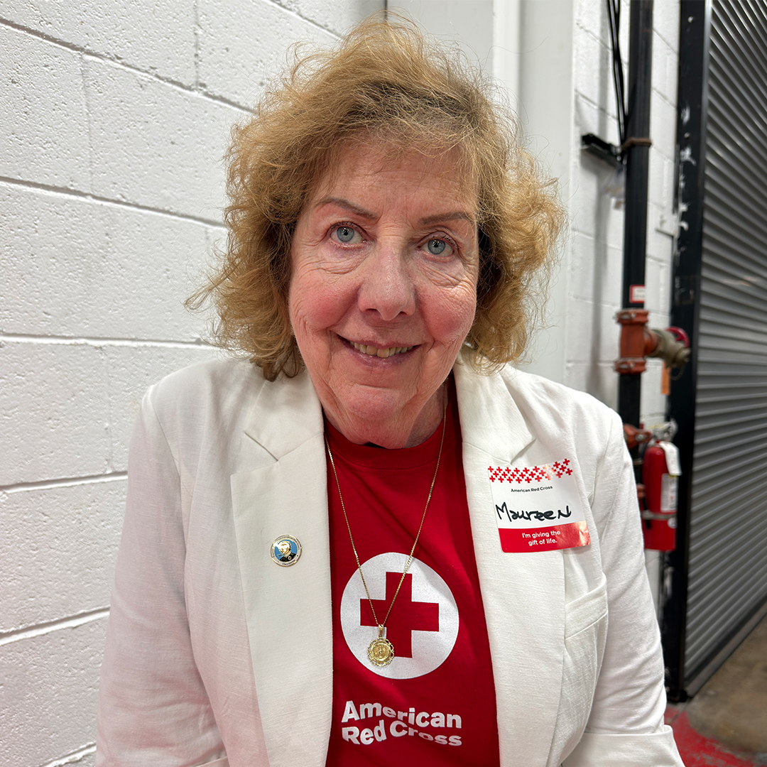 A headshot of Maureen O'Connell, a smiling woman with short, curly blonde hair and blue eyes, wearing a white blazer over a red shirt. She is wearing a Red Cross pin on her lapel and a handwritten nametag that reads "Maureen."