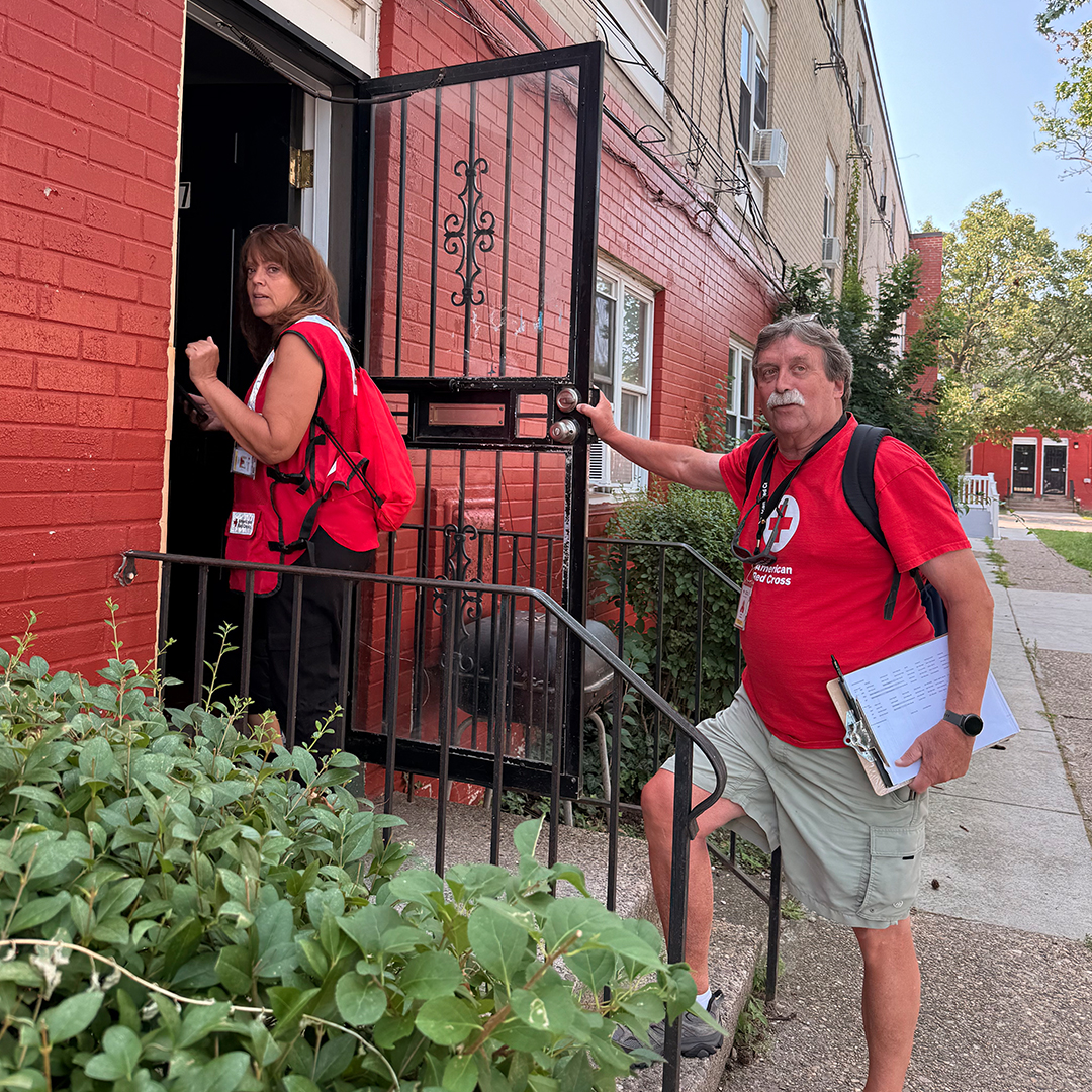 American Red Cross volunteers Griff Schrack and Linda Szypula wearing red vests, standing on the brick steps outside a residential home during a storm response.