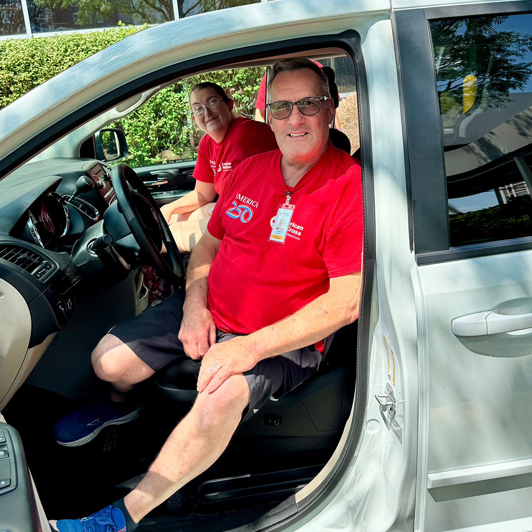 A close-up shot of two smiling American Red Cross volunteers wearing red shirts inside a white vehicle. The male volunteer in the driver's seat wears glasses and a badge, while a female volunteer sits behind him in the passenger area.