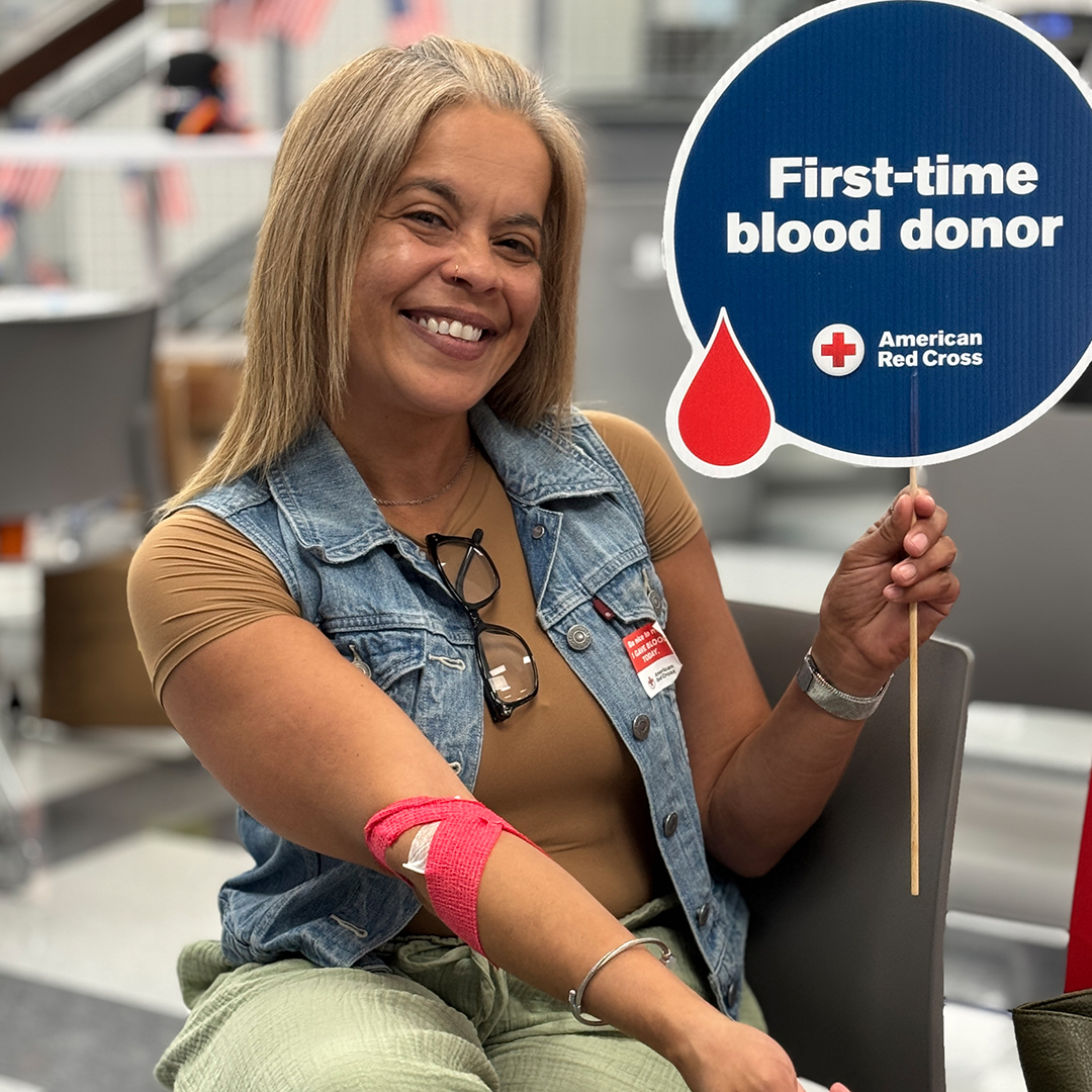 A smiling woman holds a "First-time blood donor" sign and shows her bandaged arm after giving blood.