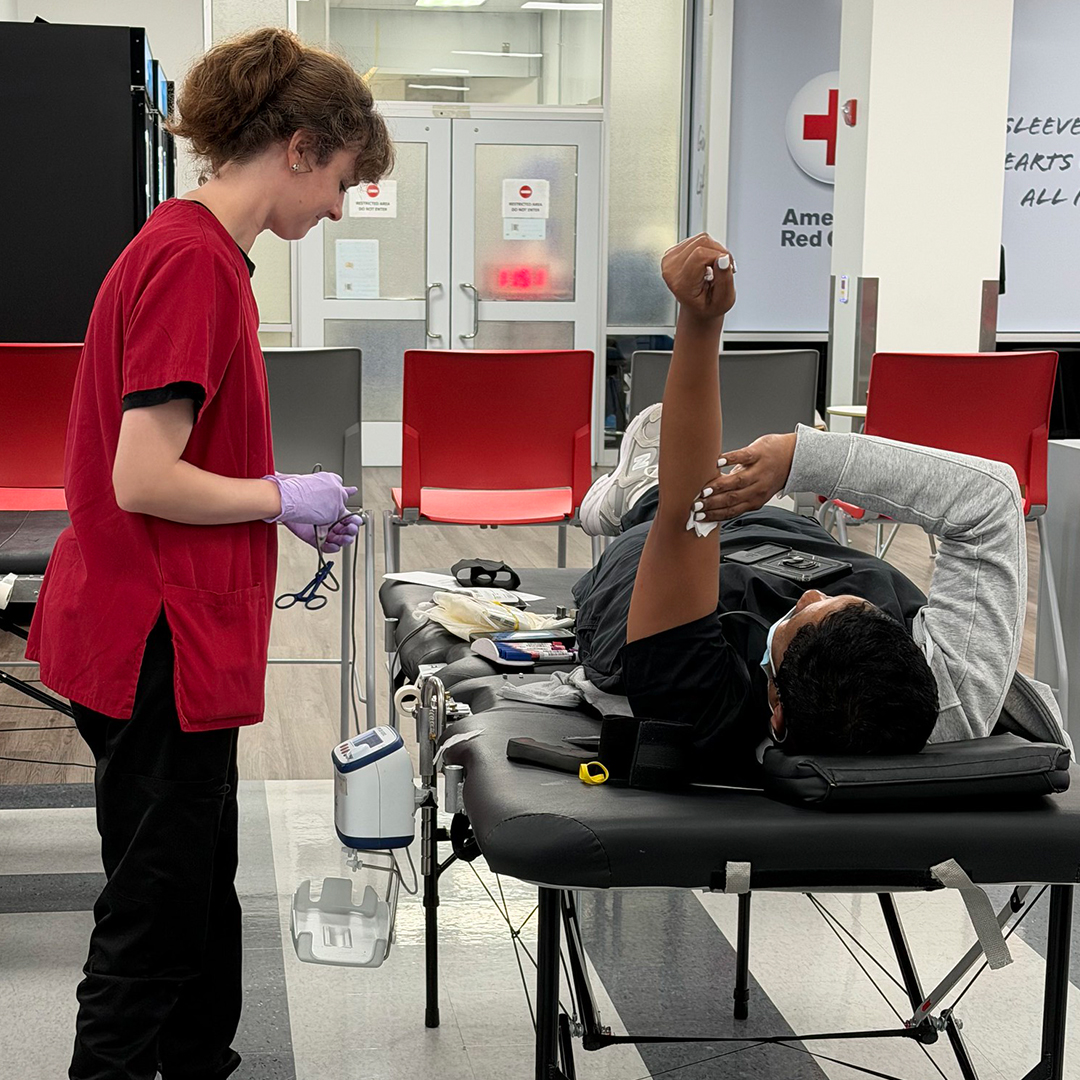 A phlebotomist in red scrubs attends to a donor lying on a table during a blood drive.