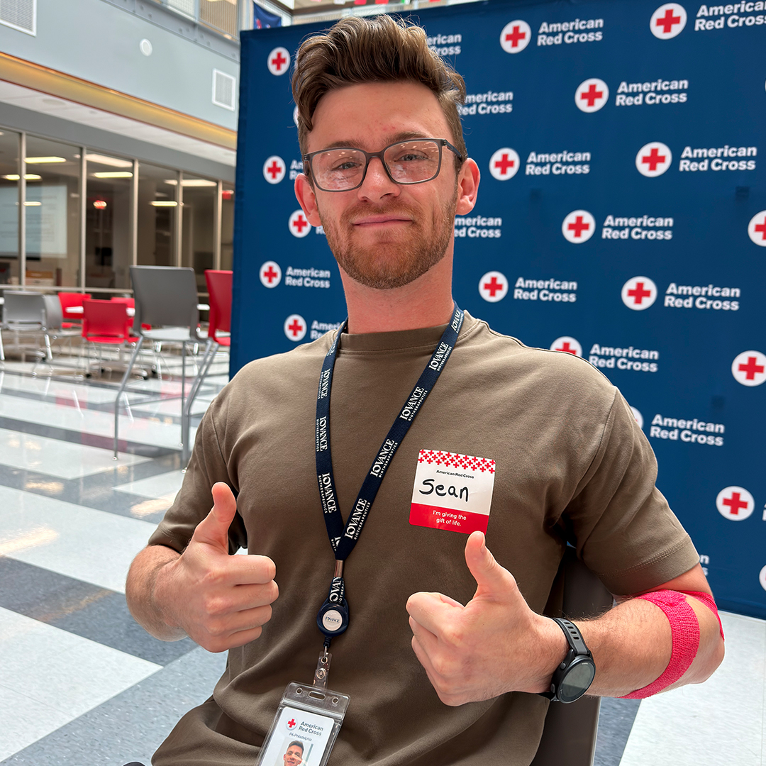 A smiling man giving a thumbs-up in front of an American Red Cross backdrop after his blood donation.