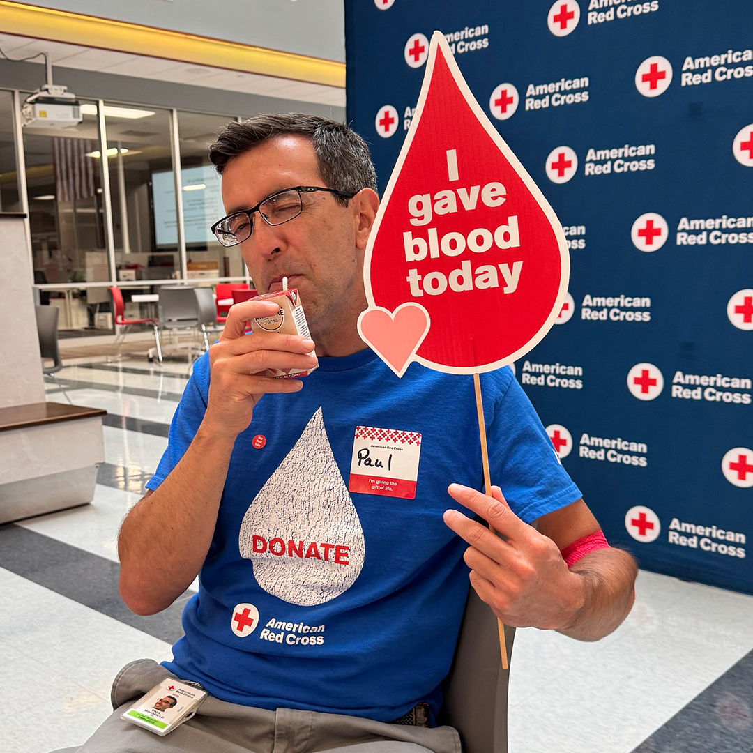A smiling man in a blue "DONATE" shirt holds a juice box and an "I gave blood today" sign at a blood drive.