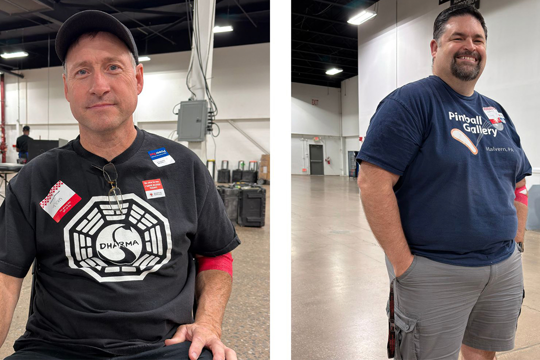 Two horizontal photos side-by-side featuring morning show super-fans. In the left photo, Tom VanDuyne sits indoors wearing a black baseball cap and a black graphic T-shirt with the "Dharma Initiative" logo from the TV show LOST. In the right photo, Craig Powell stands smiling with his hand in his pocket, wearing a navy blue T-shirt that reads "Pinball Gallery, Malvern, PA." Both men are wearing red bands on their arms from their blood donations.