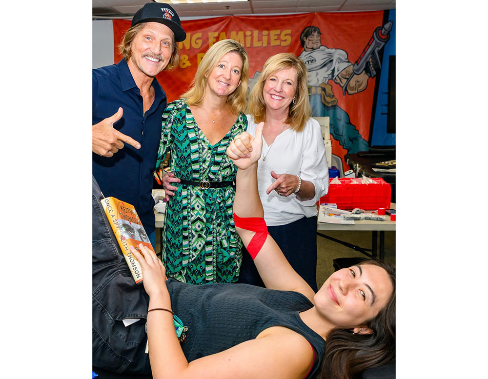 Charles Antis, founder and CEO of Antis Roofing and Waterproofing, Cori Vernam, Susan DeGrassi cheer on a blood donor who is lying on a cot giving a thumbs up.