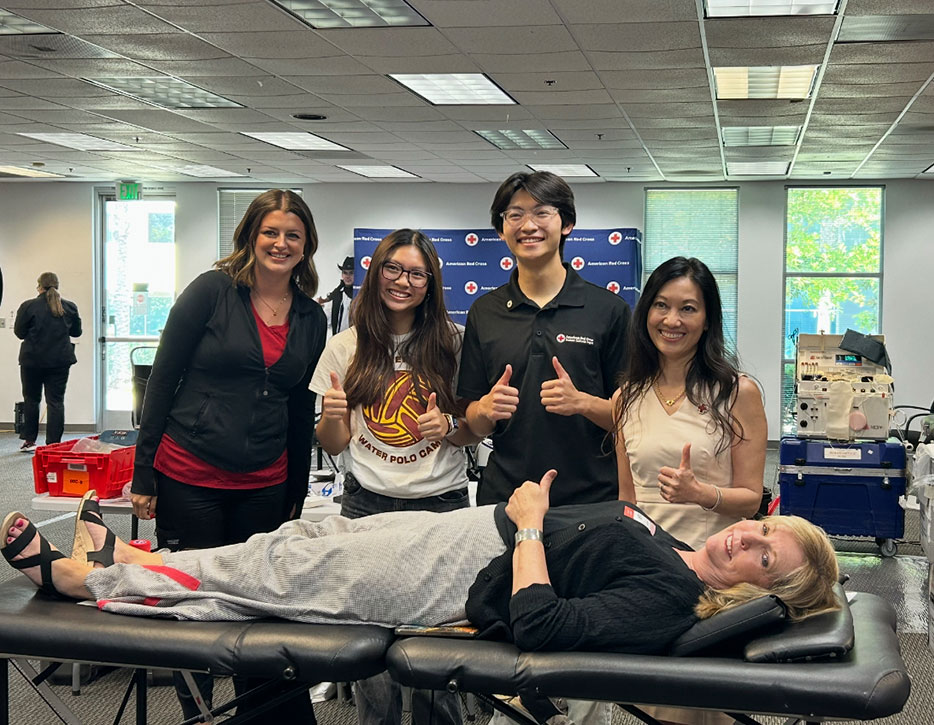 Red Cross of Orange County Board Vice Chair Susan DeGrassi lying on a cot after donating blood, with other board members standing next to her, all giving a thumbs up.