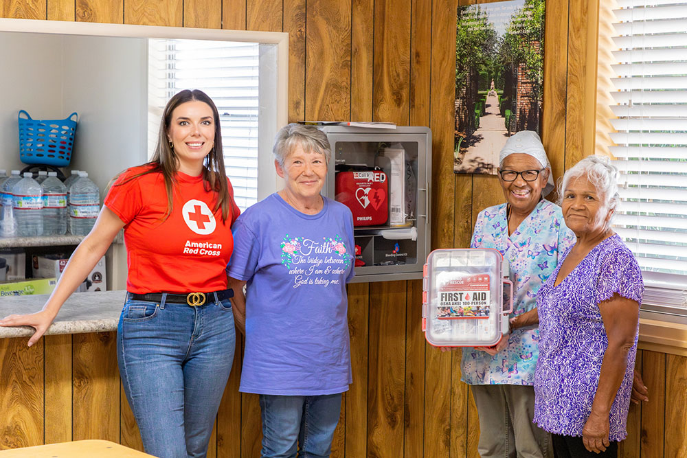 Four women stand and talk to each other in front of a counter next to an Automated External Defibrillator mounted on the wall while displaying a first aid box.