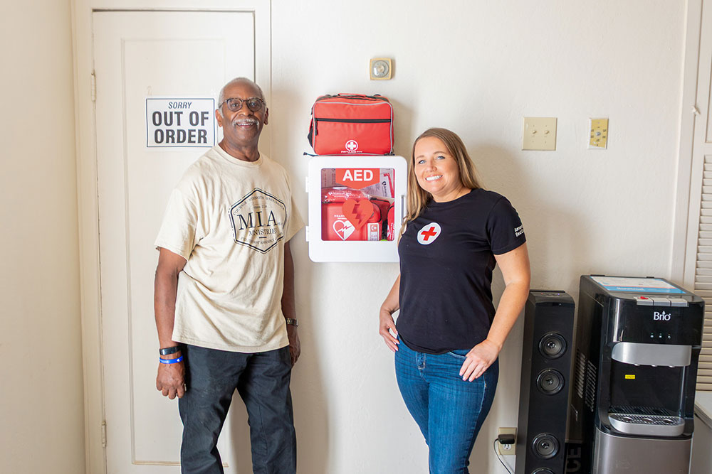Two people standing next to an Automated External Defibrillator mounted on a break room wall.