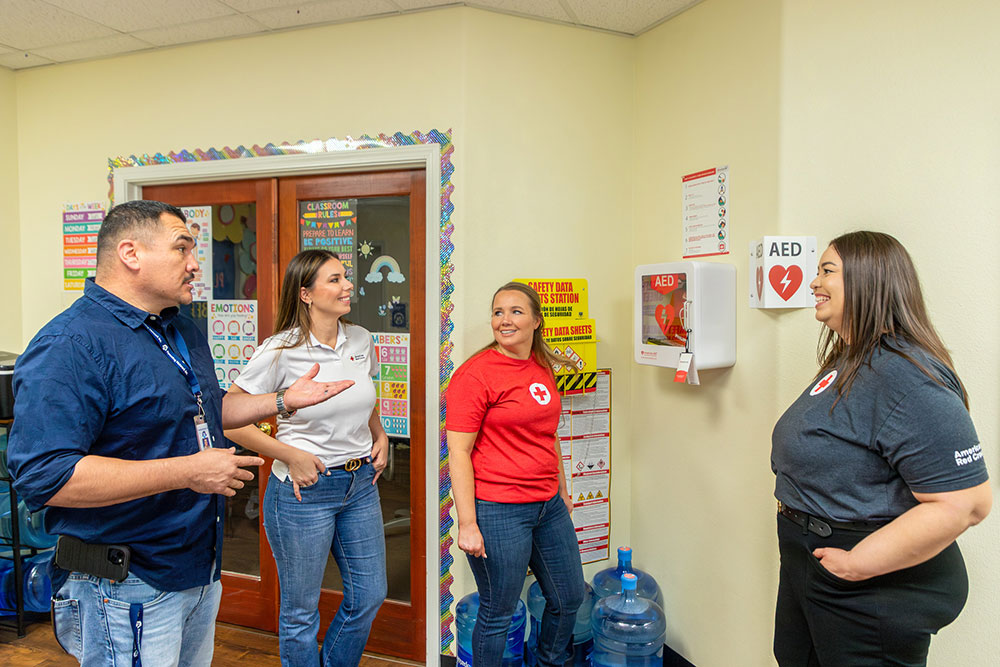 Four people talking to each other next to an Automated External Defibrillator mounted on a break room wall.