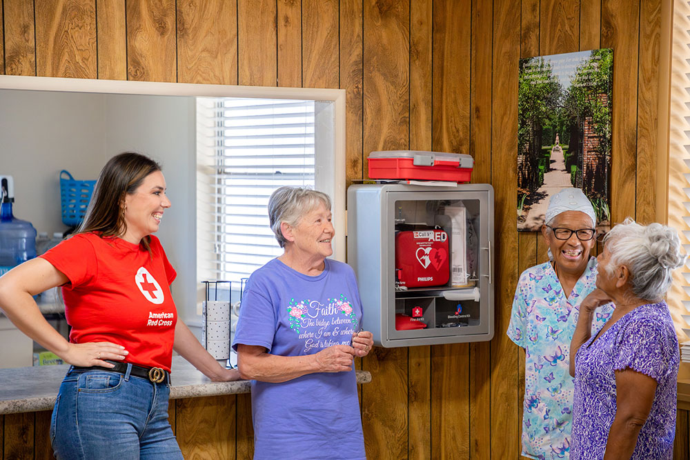 Four women stand and talk to each other in front of a counter next to an Automated External Defibrillator mounted on the wall.