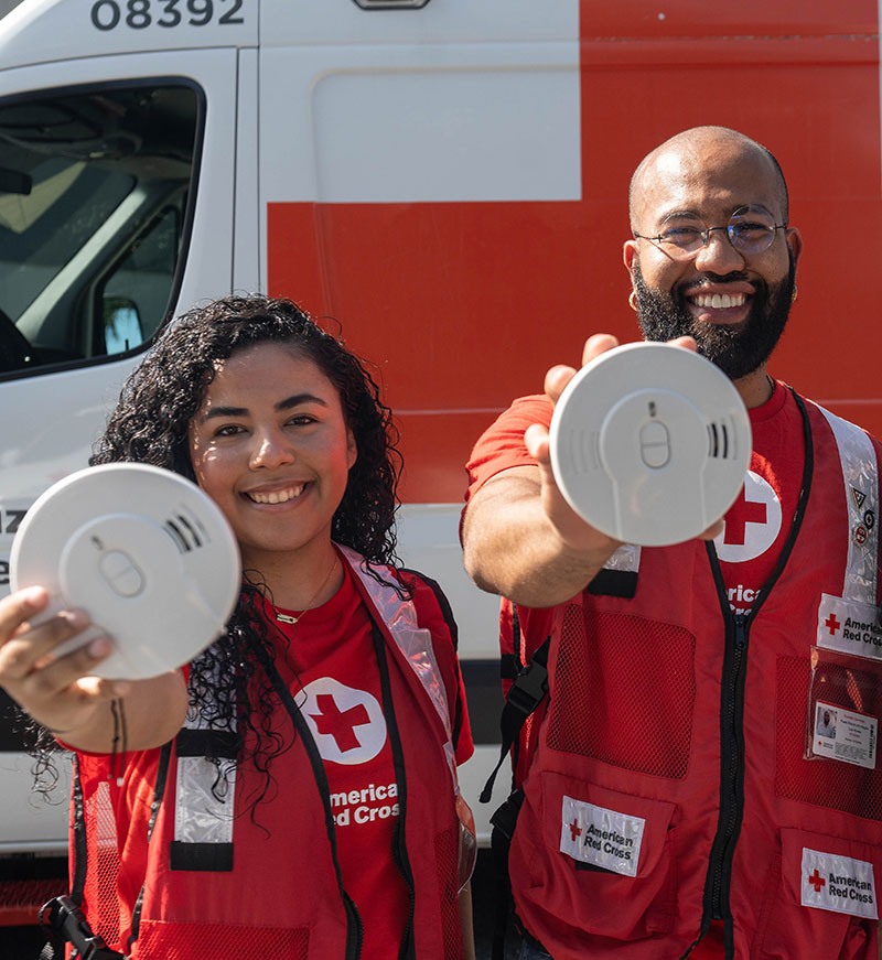 two Red Cross volunteers holding smoke alarms and smiling.