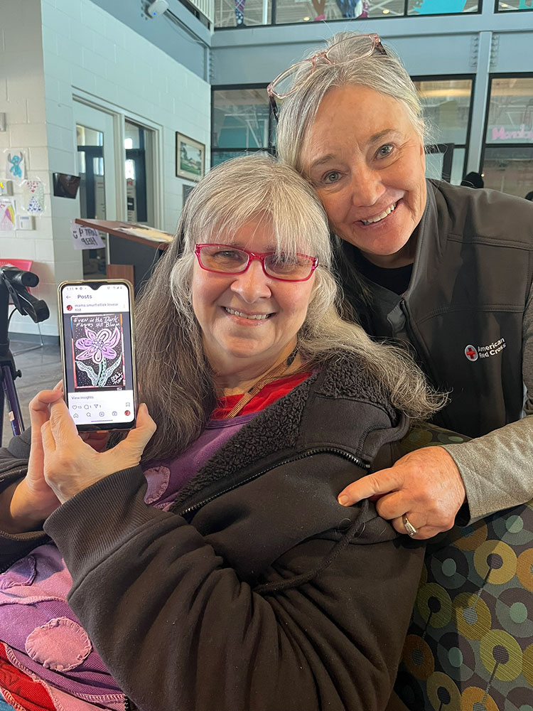 Ellie Kane, next to a Red Cross shelter volunteer, holding up a smart phone that has a picture of a sketch she made.