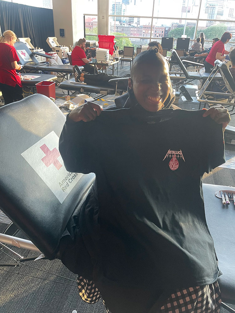 Jamilah Bourdon holding up a Red Cross x Metallica Blood Drive T-Shirt while sitting on a blood donation chair. 