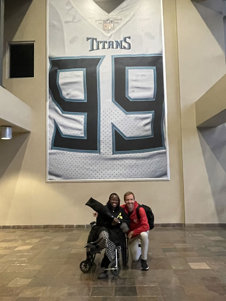 Jamilah Bourdon in wheelchair with another person kneeling with arm around Jamilah and a large Tennessee Titans jersey picture hanging in the background.
