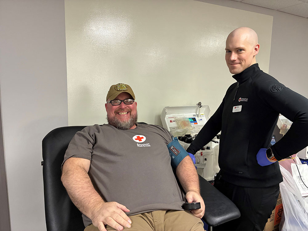 Joseph Hall sitting in a blood donation chair, wearing a Red Cross shirt. A Red Cross staff member stands beside him.