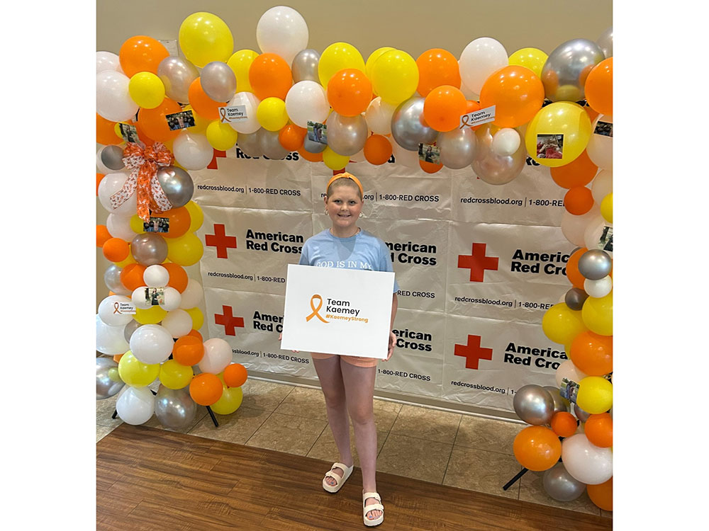 Kaemey Young holding a Team Kaemey sign with balloons and American Red Cross signs behind her.