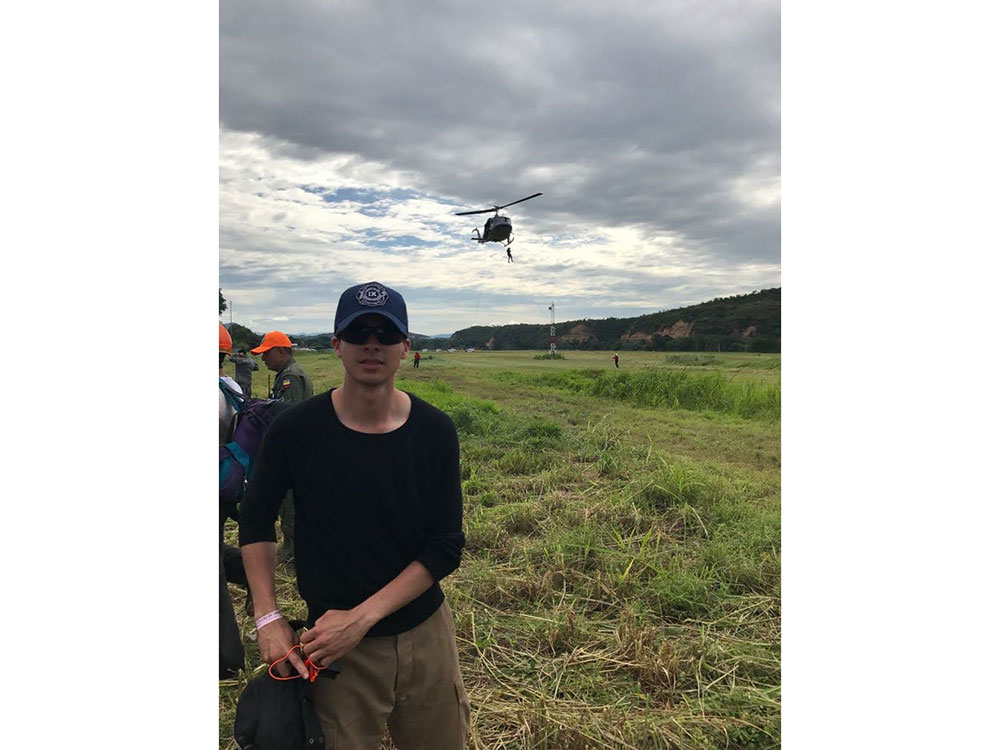 Camilo Riviere wearing a hat and sunglasses in an open field of grass with a helicopter in the background and a person rapelling off it.