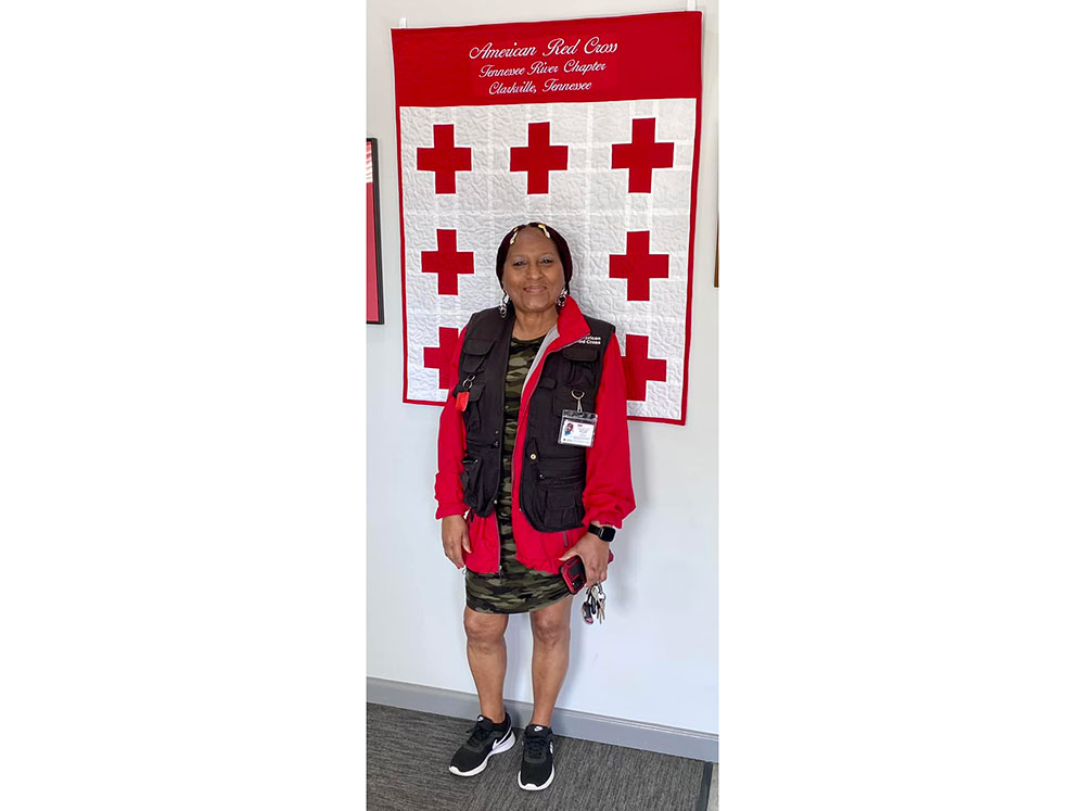 Red Cross volunteer Mary Lockett in front of a Red Cross quilt hanging on a wall.