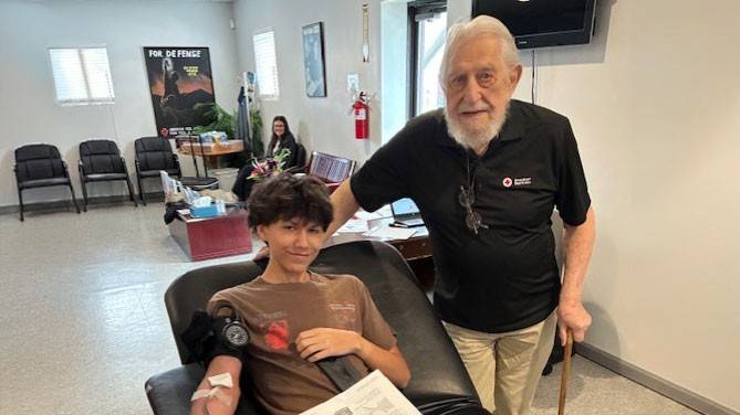 Ray Swadley’s grandson donates blood, lying on a reclined chair, smiling and holding a stress ball. Ray Swadley stands beside, smiling supportively.