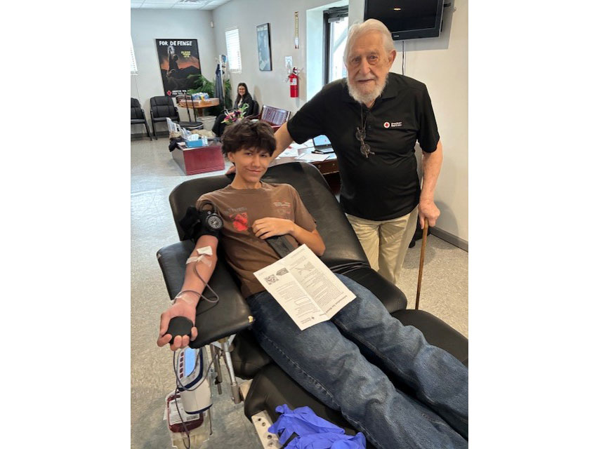 Ray Swadley’s grandson donates blood, lying on a reclined chair, smiling and holding a stress ball. Ray Swadley stands beside, smiling supportively.