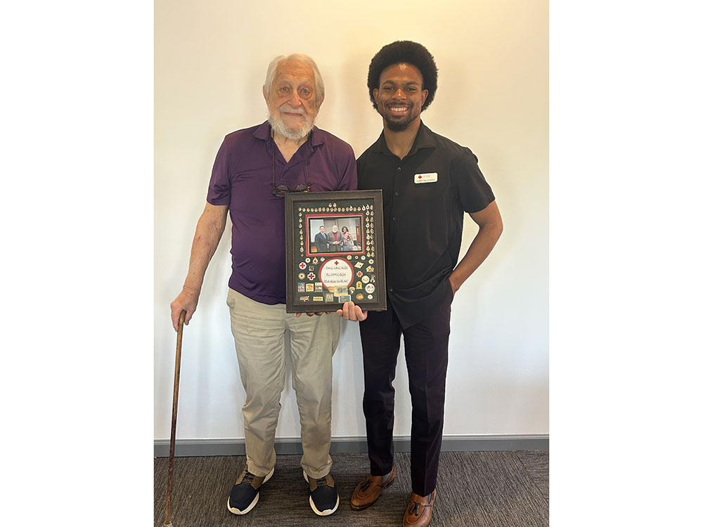 Ray Swadley, holding a cane, a framed photo and memorabilia, while standing next to a Red Cross worker.