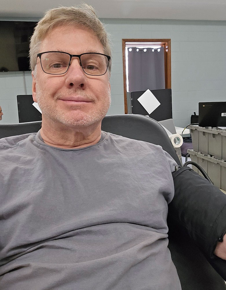 Stephen Kinzer wearing glasses sits in a chair and donates blood.