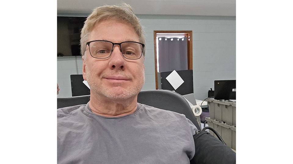 Stephen Kinzer wearing glasses sits in a chair and donates blood.
