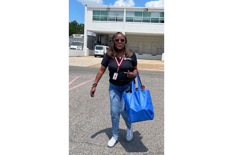 Tena Wheat outside wearing shades, a red cross shirt and lanyard, while carrying reusable shopping bags.