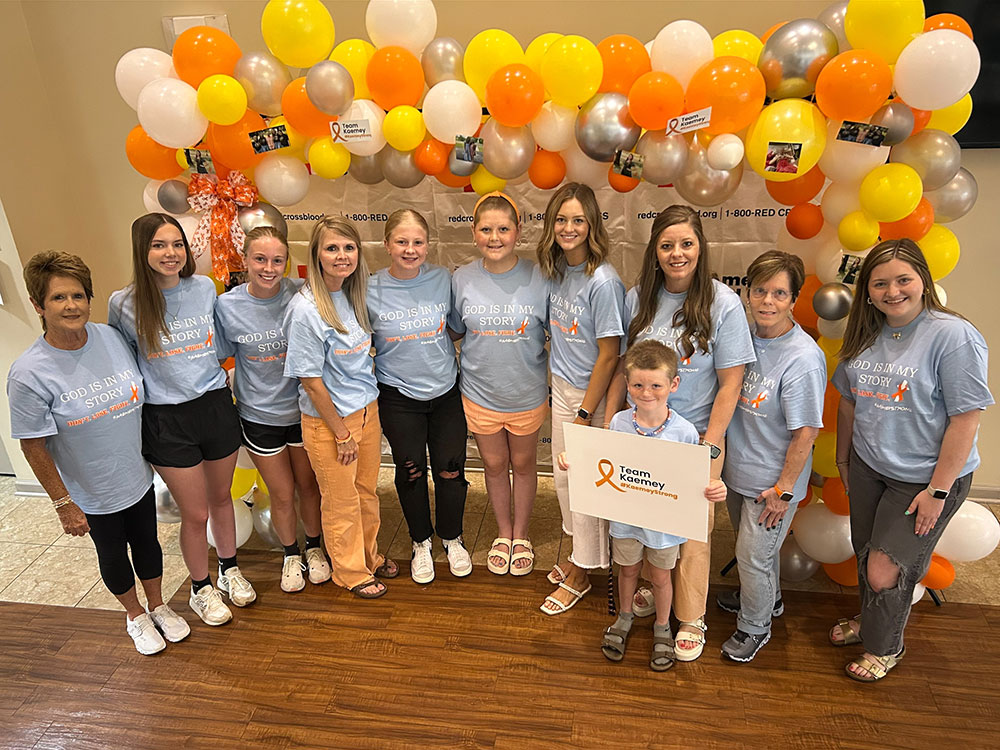Kaemey Young and a group of blood donors with one of them holding a Team Kaemey sign with balloons and American Red Cross signs behind them.