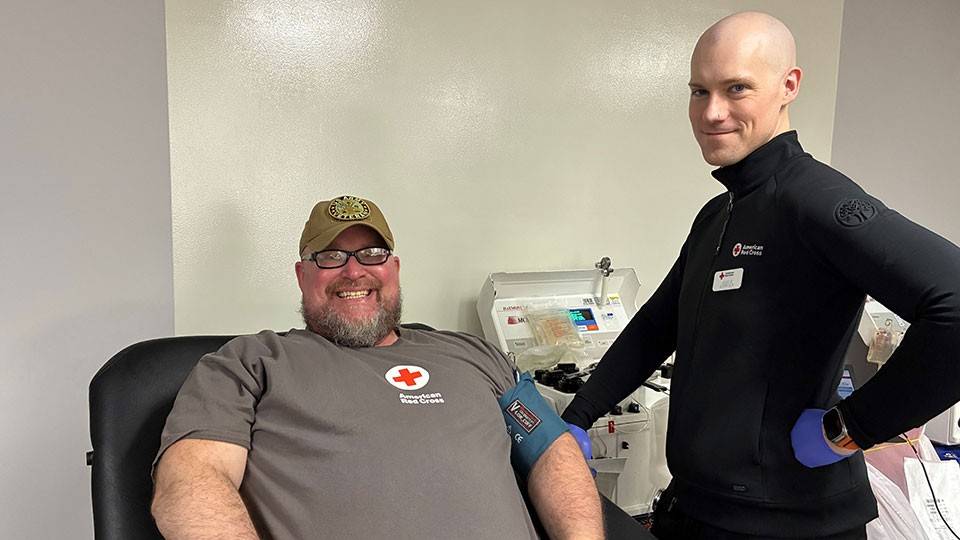 Joseph Hall sitting in a blood donation chair, wearing a Red Cross shirt. A Red Cross staff member stands beside him.